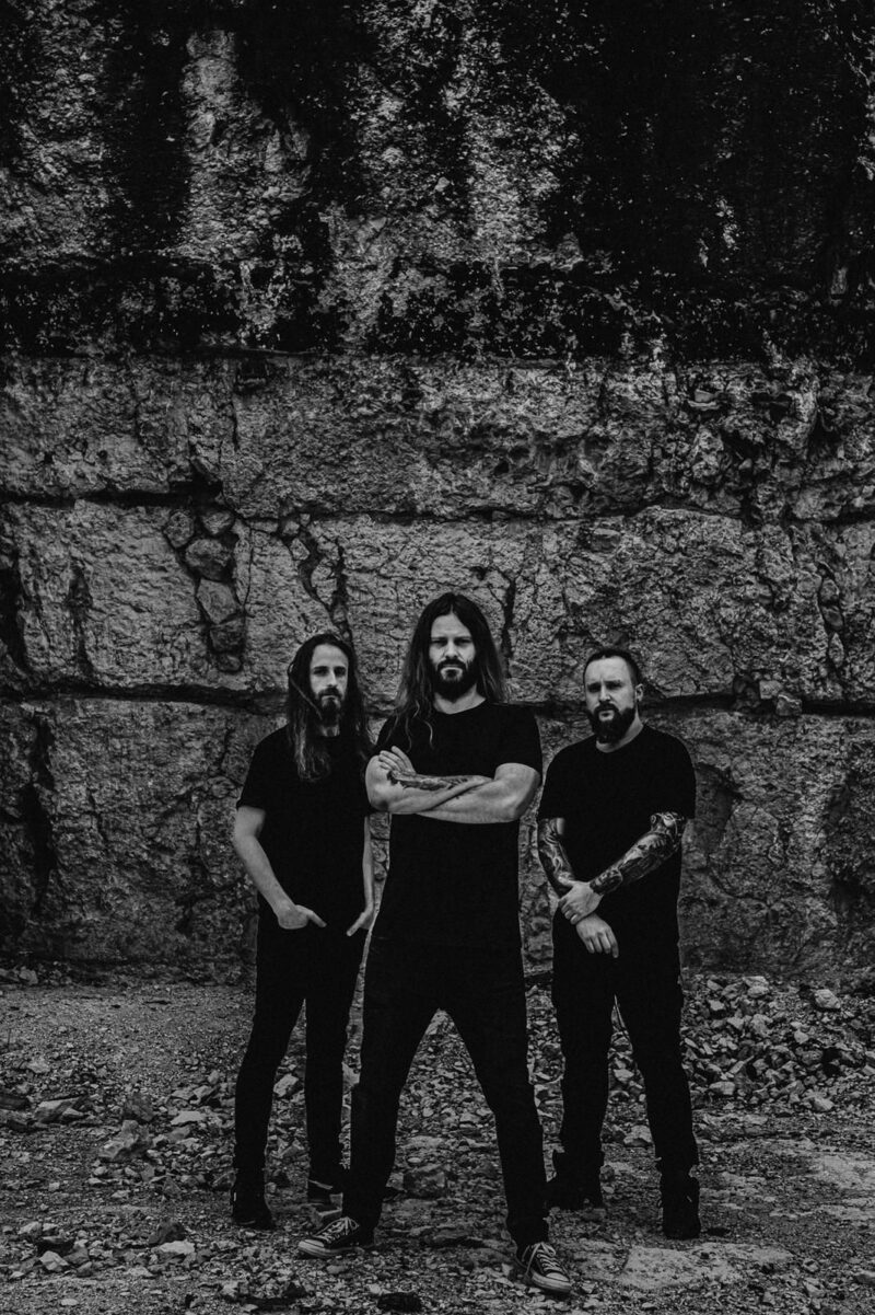 Svartvit bild på tre medlemmar i bandet Decapitated. De står framför en stenvägg.