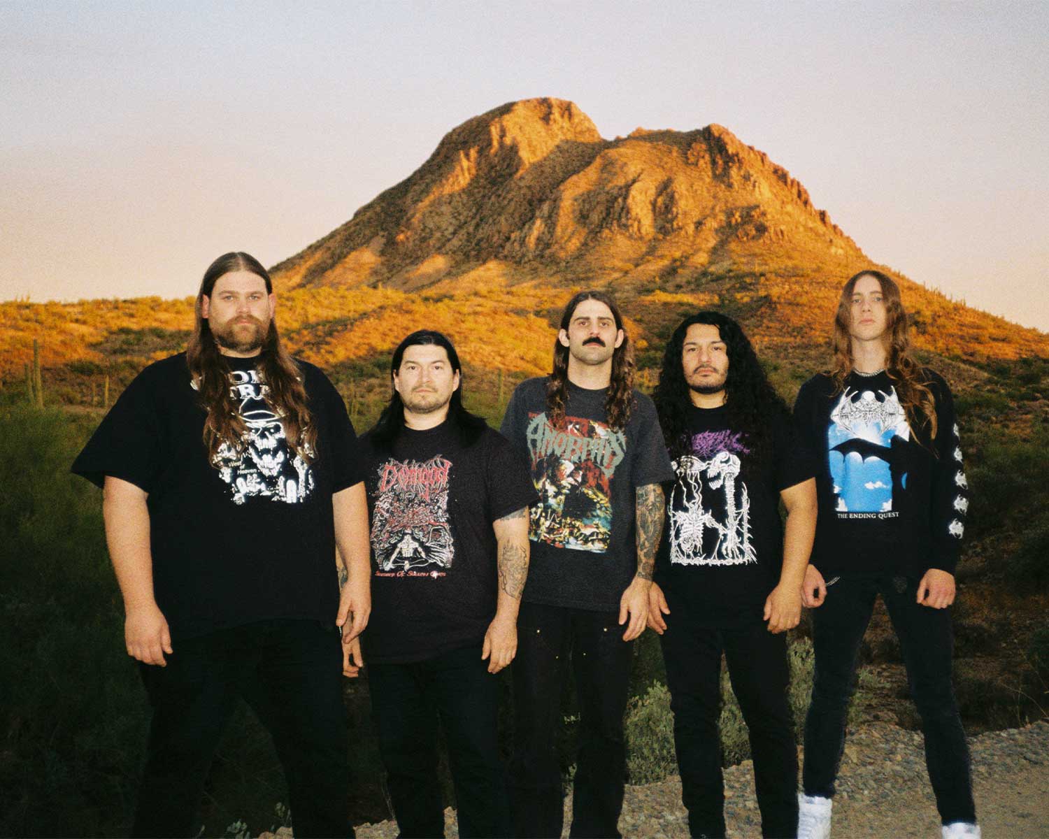 Fem bandmedlemmar från Gatecreeper kollar in i kameran, klädda i band t-shirts och svarta byxor. Bakom personer syns ett högt berg i orange färg.