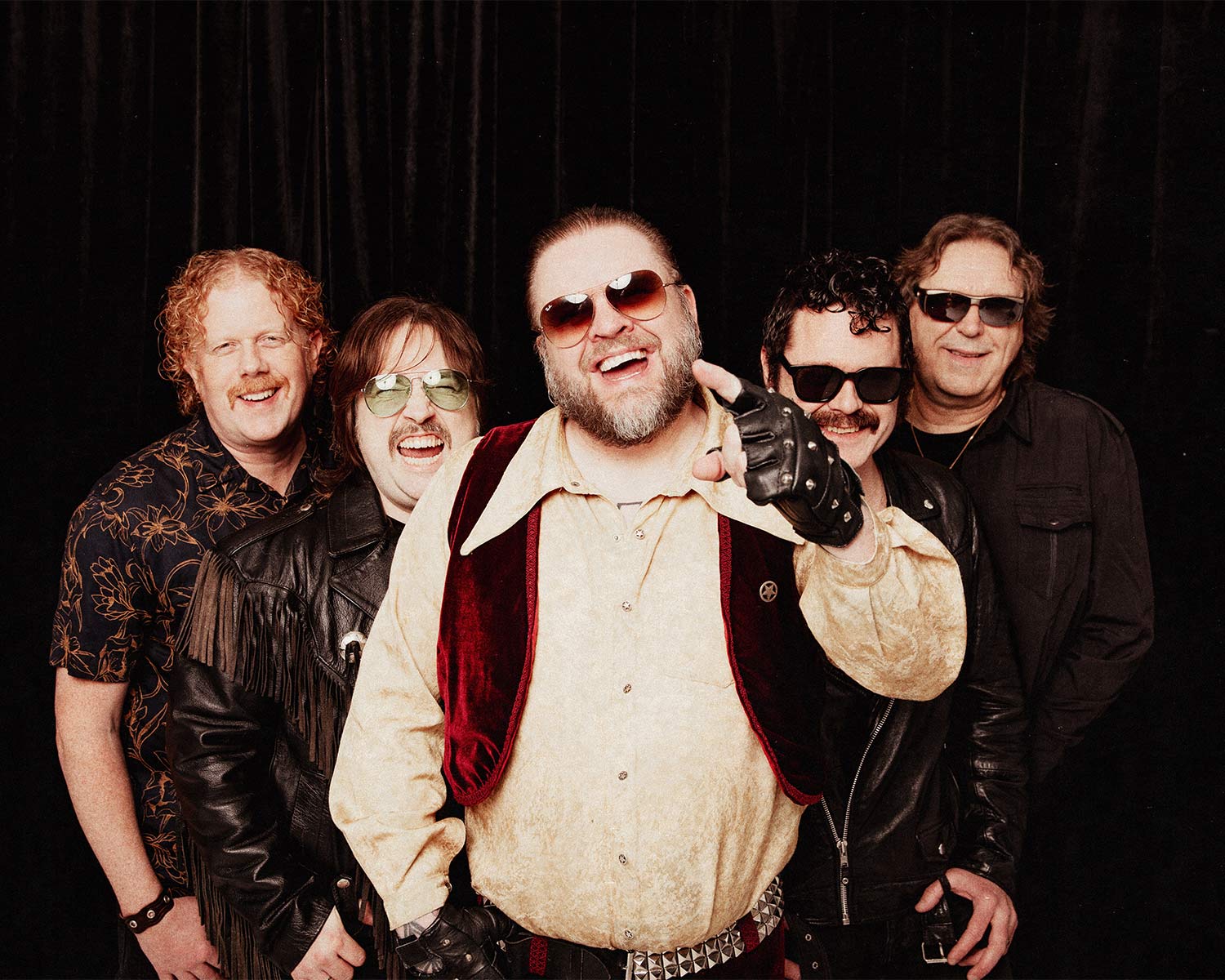 Pressbild av Korslagda: fem bandmedlemmar poserar tätt tillsammans framför svart bakgrund. Personen i mitten pekar mot kameran och bär röd väst samt solglasögon, övriga medlemmar har skinnjackor och solglasögon i en avslappnad, humoristisk rockpose.