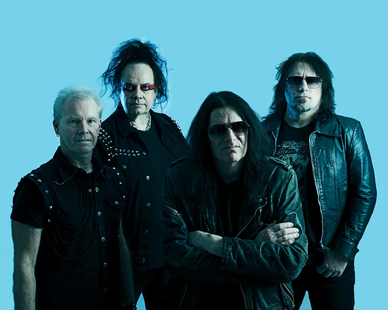 Pressbild av Lion’s Share: fyra bandmedlemmar poserar framför blå bakgrund. De bär svarta kläder och skinnjackor, och en medlem har mörk sminkning runt ögonen medan de andra står bakom och tittar mot kameran.