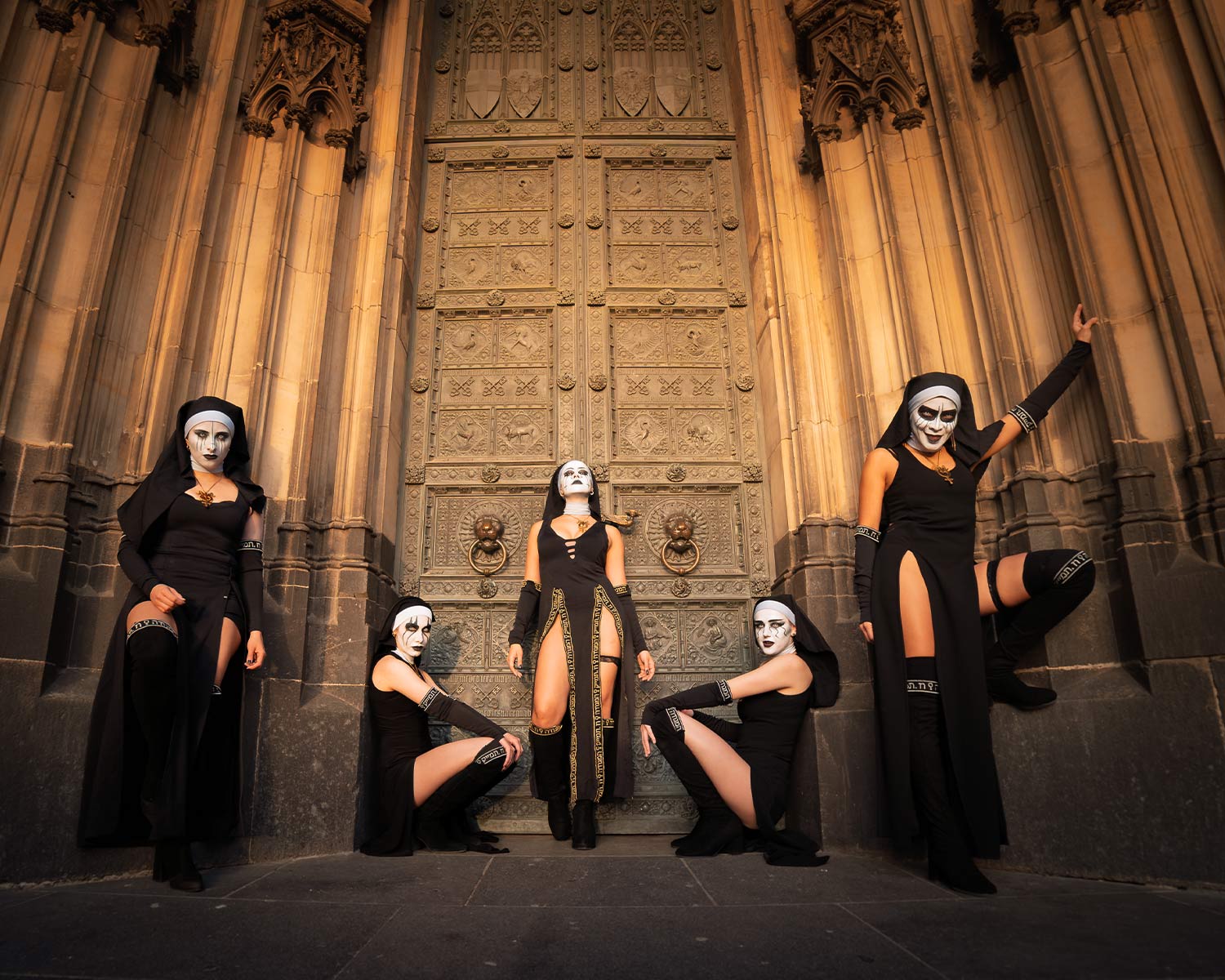 The members of Dogma pose in black, nun-inspired stage costumes in front of a large church gate.