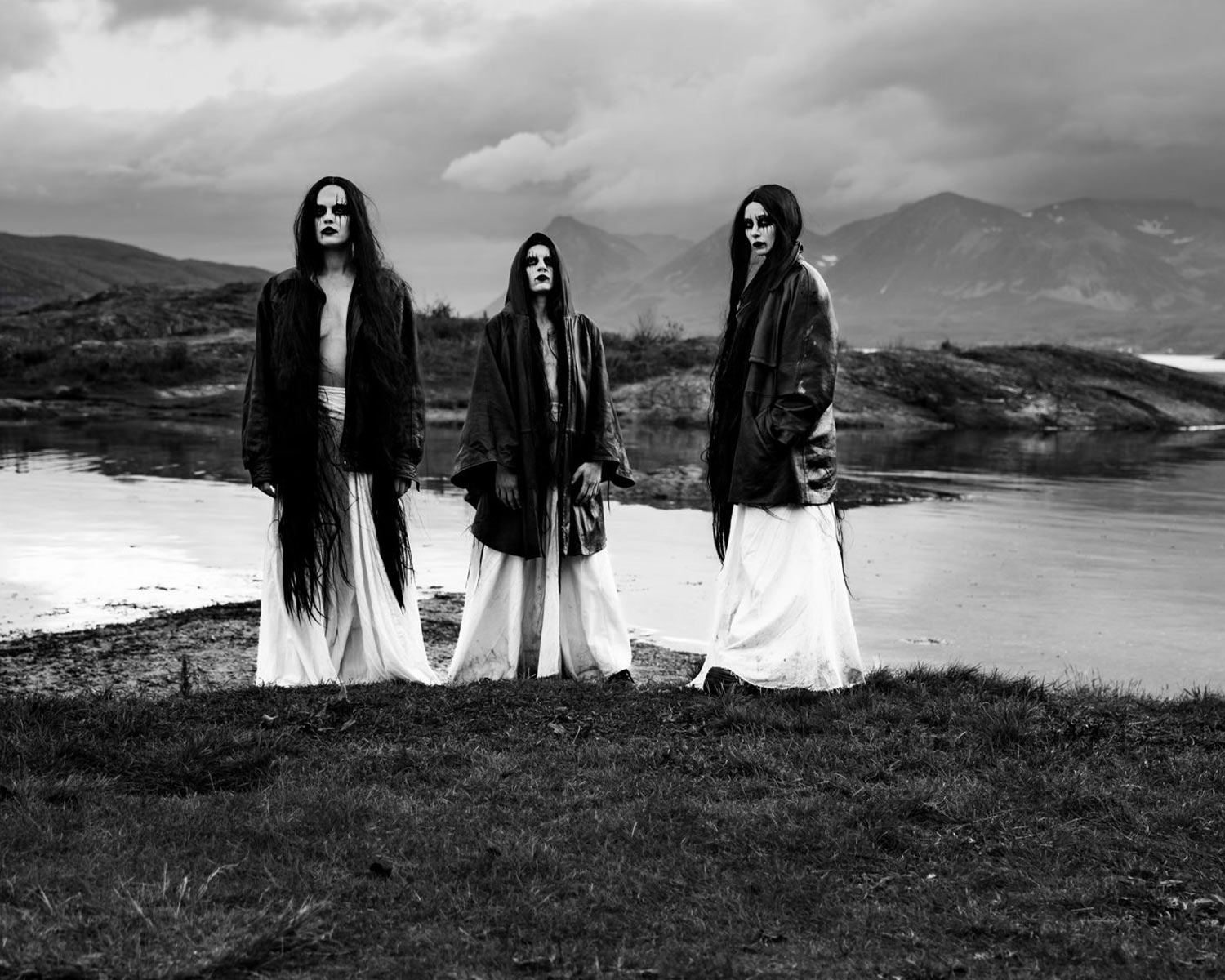 The members of the Witch Club Satan are standing by a lake in black-and-white, dressed in dark cloaks and white dresses.