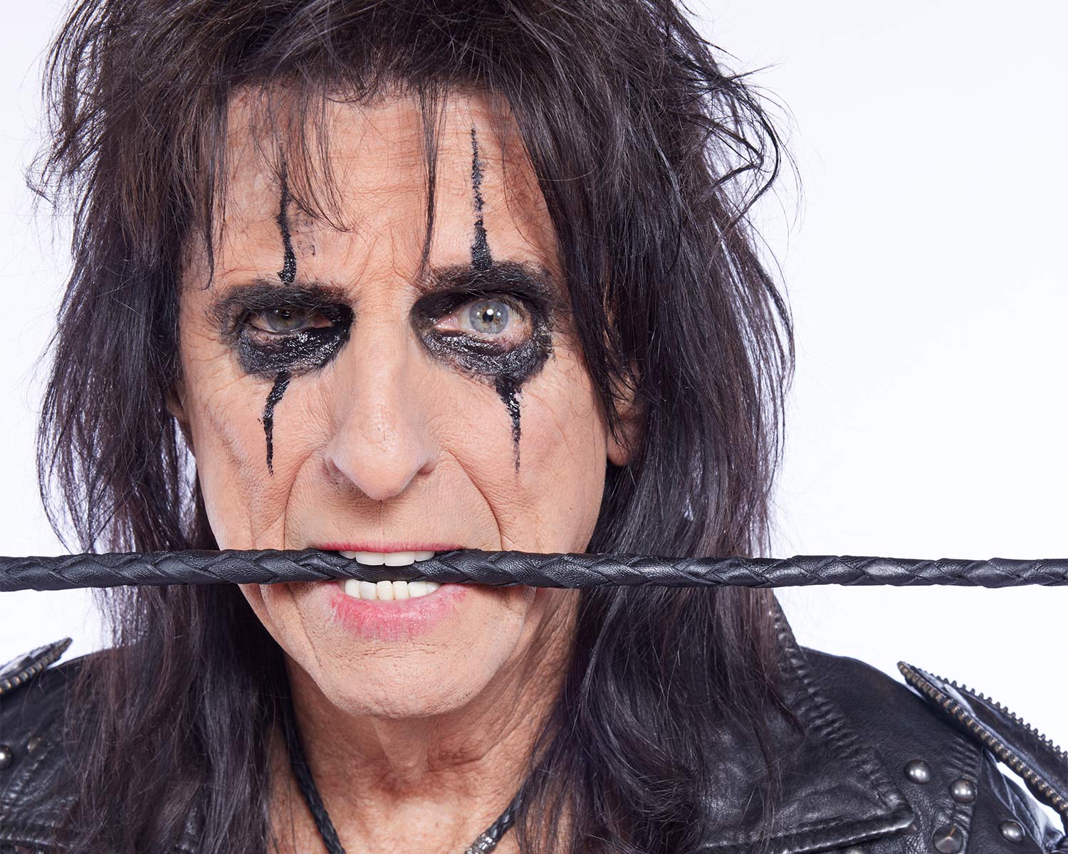 A close-up of Alice Cooper, wearing black makeup around his eyes and holding a leather whip in his mouth.