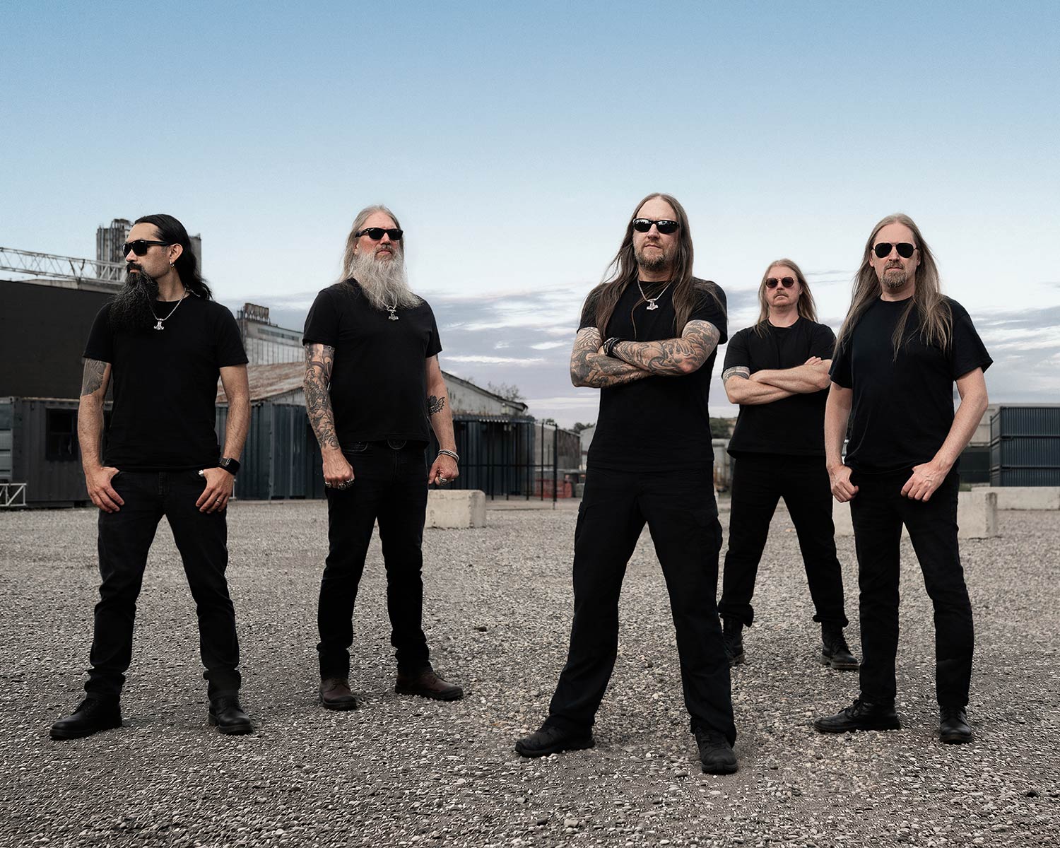 The members of Amon Amarth pose in black clothing at an industrial yard.