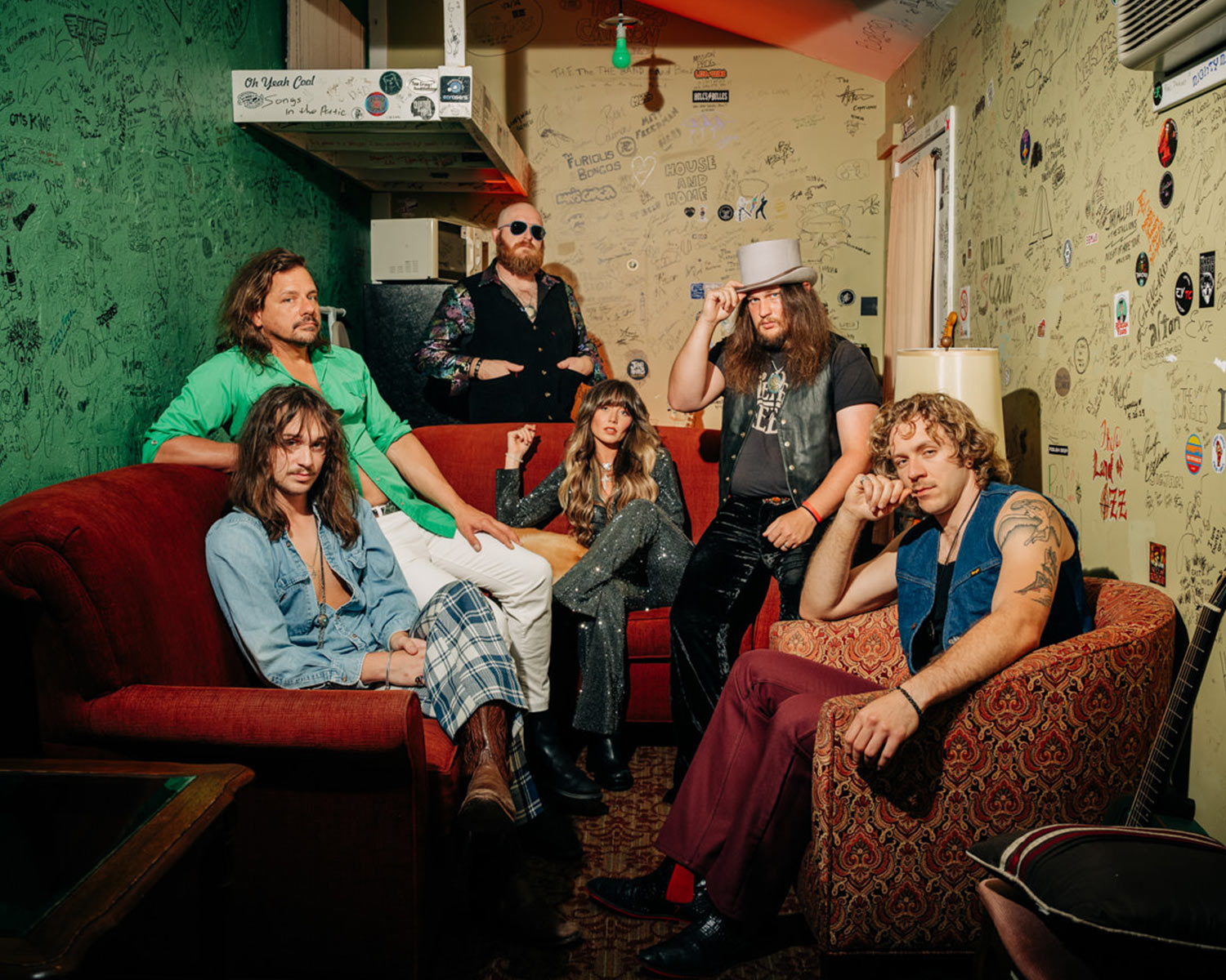 Six band members are sitting on red sofas and armchairs in a room with green and yellow walls.