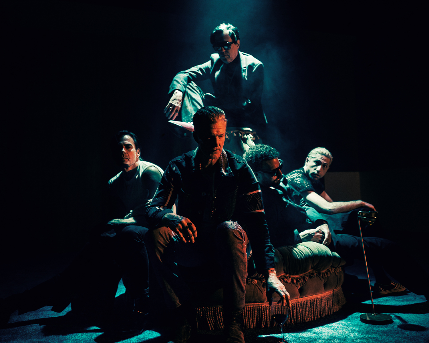 The five members of Queens of the Stone Age pose in dim blue-green lighting, sitting on and around a sofa.